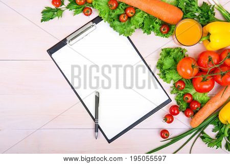 A close-up white sheets of papers with a pen. Different veggies on a bright wooden background. Delicious vegetables. Green lettuce, onions and parsleu. Fresh red tomatoes and carrots.