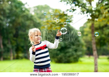 Little blonde cheerful girl playing with soap bubbles in the green summer park. Outoors.