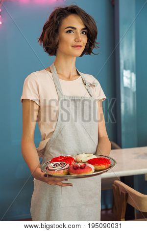 Portrait of a young pretty waitress in apron serving donuts on a tray in a caffeteria