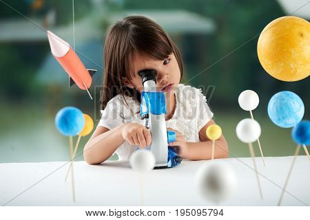 Curious little girl looking through microscope while having fun in scientific club for preschoolers, blurred background