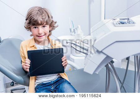 Portrait Of Little Boy Sitting On Chair And Showing Tablet In Hands In Dental Clinic