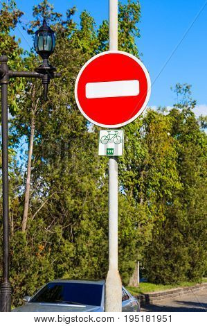 Round red No Entry road sign mounted on gray metal pole, green trees and grey car background. With bycicle allowed route square sign.