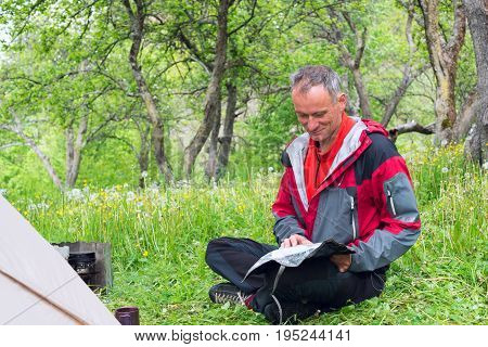 Smiling Man Traveler Sits On A Green Meadow, Next To His Tent