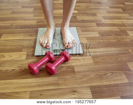 Female legs stand on the scales and two red dumbbells lie near on the floor. Two slender legs with a red pedicure gray electronic scales and two red dumbbells lie on the brown floor