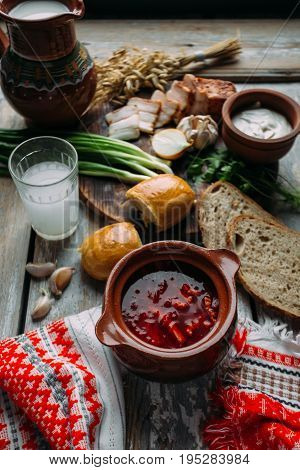 Borsch - beetroot soup in a clay bowl on a wooden background, traditional dish of ukrainian and russian cuisine. In the background there is a cutting of bacon, gorilka, green onions, garlic