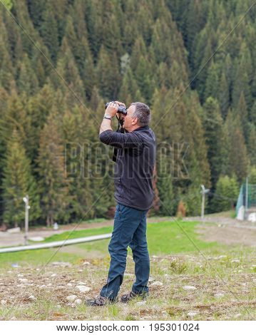 Romanian Tourist With Binocular In Hands Stand On A Cliff And Watching Into Deep Misty Valley Bellow The Retezat Mountains