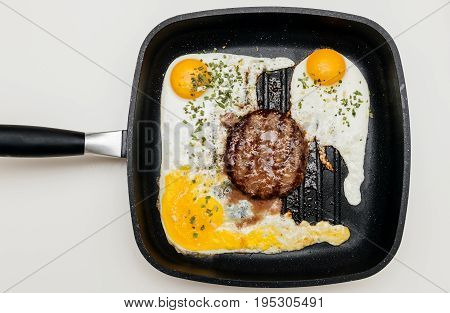 Overhead view above of delicious raw beef meat burger in a frying pan on cooking surface in kitchen with three fried eggs and one broken yolk