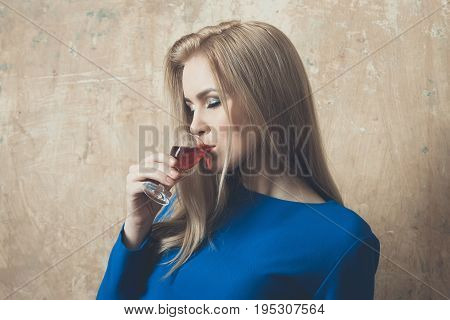 Girl enjoying red liqueur in glass. Woman with blond long hair make up in blue dress on beige wall. Alcohol appetizer bad habits addictive and convive. Unhealthy lifestyle copy space