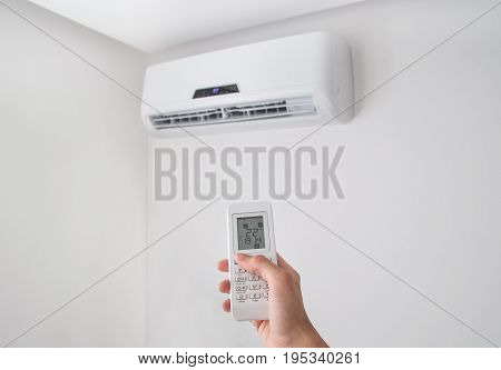 Hand holding remote control, adjusting temperature of air conditioner mounted on a white wall. Indooor comfort temperature