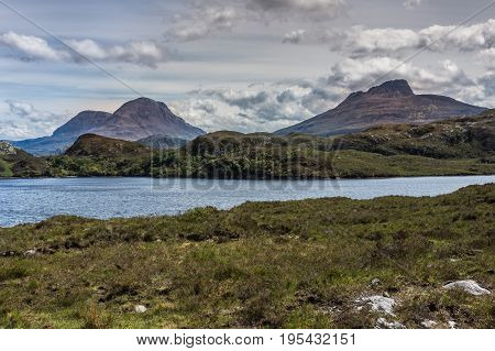 Assynt Peninsula Scotland - June 7 2012: Three brown mountain peaks under heavy brownish skies fronted by green wild rough slopes east of Loch Buine Moire. Lake waters in front.