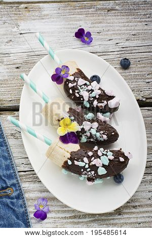 Chocobanana Light summer refreshing dessert made from frozen banana Eskimo in a glaze made of dark chocolate and colored meringue on a cocktail tube on a light background. Top View.