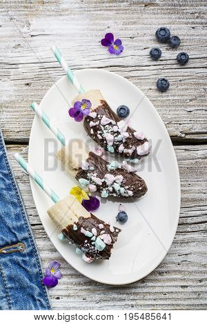 Chocobanana Light summer refreshing dessert made from frozen banana Eskimo in a glaze made of dark chocolate and colored meringue on a cocktail tube on a light background. Top View.