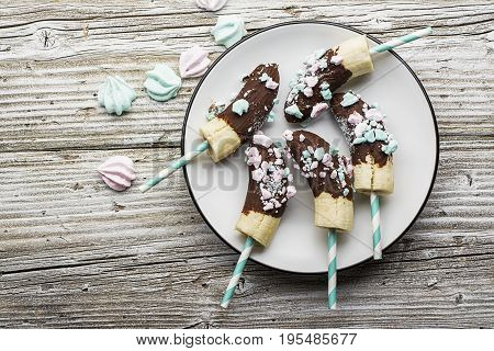 Chocobanana Light summer refreshing dessert made from frozen banana Eskimo in a glaze made of dark chocolate and colored meringue on a cocktail tube on a light background. Top View.