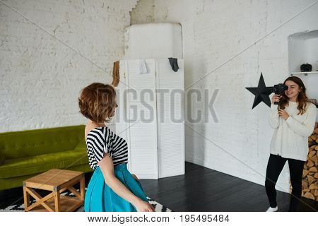 Candid shot of female photographer in white sweater holding camera taking pictures of happy young woman in beautiful clothing during fashion photo shoot in professional photographic studio interior