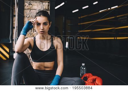 Beautiful sporty girl looking at camera and sitting on the floor in the gym after hard workout. She is holding up her head with right arm because of tiredness