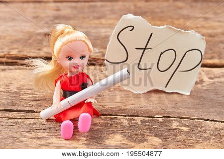 Toy doll holds a cigarette. Doll with cigarette, message stop, old wooden background. Metaphor concept stop smoking.