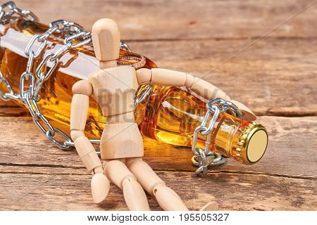 Human wooden dummy sitting with bottle. Bottle wrapped in chain, wooden human mannequin, old wooden background. Concept of alcohol addiction.