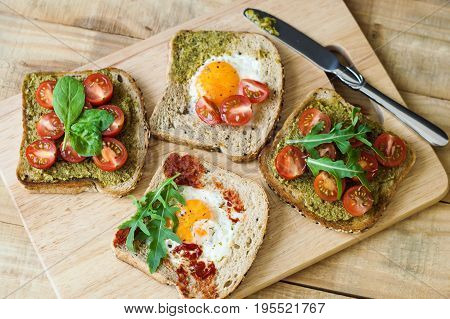 Breakfast toasts on wooden cutting board. Healthy sandwiches with cherry tomato egg spinach arugula pesto sauce tomato sauce and bread with sesame seeds. Balanced breakfast