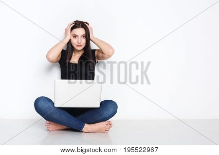 Schocking news. Worried woman sitting at home on floor and looking on screen of laptop while holding hands on her head.