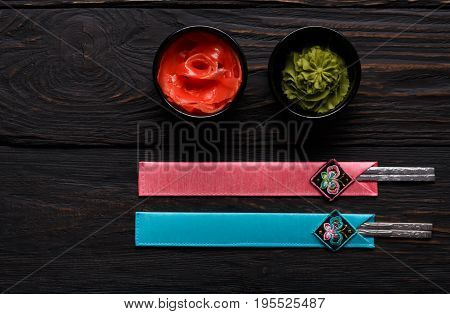 Pieces of traditional japanese tablewear. Chopsticks in blue and pink cases, small black bowls with ginger and wasabi on dark wooden table. Top view, flat lay, copy space, background