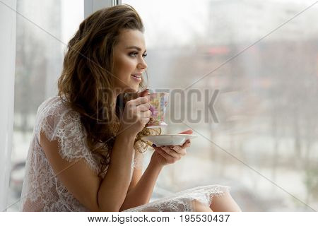 Sexy Young Woman In Erotic Transparent Underwear Drinks Coffee On Window Sill. Cityscape On Backgrou