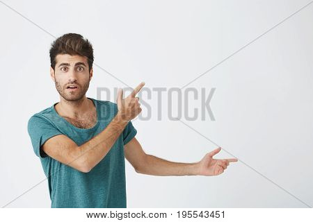 Indoor shot of astonished good-looking man dressed in casual cloth having excited fascinated look, indicating at blank studio wall with his index finger. Isolated shot, horizontal.