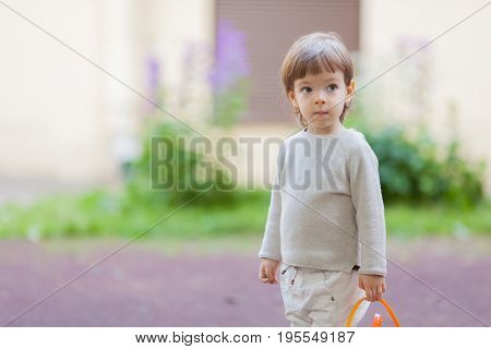 Little Beautiful Boy With Long Hair And Blue Eyes Holding A Bucket Of Toys For The Sandbox