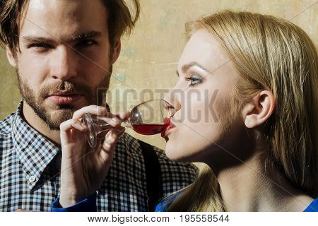Girlfriend drinking red liqueur in glass and boyfriend. Woman or girl with blond hair and handsome man or macho on beige wall. Couple in love. Alcohol and convive. Unhealthy lifestyle. Bad habits