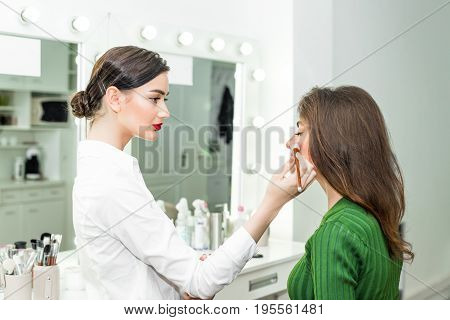 Attractive brunette girl primp at the mirror with lamps in the beauty studio. A happy satisfied customer of make-up and hairstyle service.