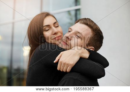 Happy beautiful young Caucasian couple cuddling outdoors. Beautiful brunette woman closing eyes in pleasure while hugging her bearded boyfriend after long separation. Love and happiness concept