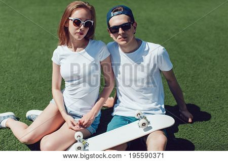 Young skater boy and girl with skateboard in white T-shirts are sitting on the lawn. Mock up.