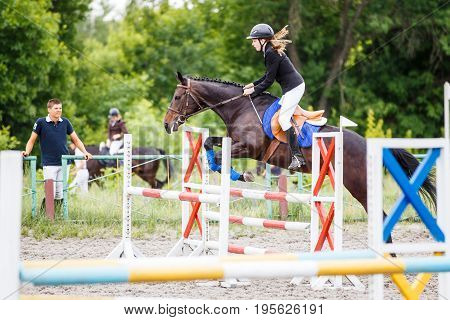 Young rider girl on bay horse training jumping over obstacle with her trainer