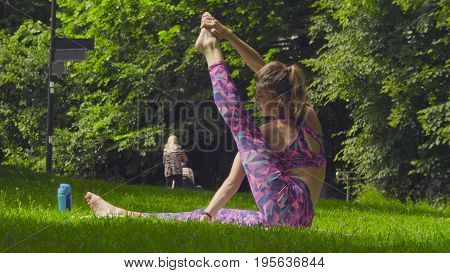 Young woman doing yoga exercises in the park. Akarna Dhanurasana