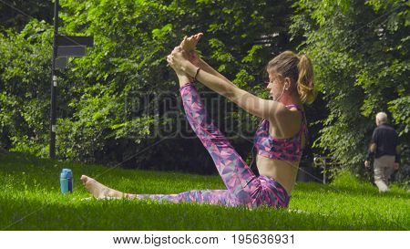 Young woman doing yoga exercises in the park. Akarna Dhanurasana