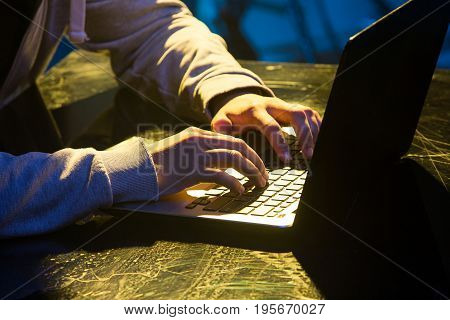 Hooded computer hacker stealing information with laptop on colored studio background