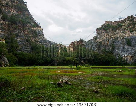 Wide-angle view of the curved craggy peaks of Khao Ngu Stone Park with a grassy wetland and trees in the foreground. Ratchaburi Thailand. Travel and environment concept.