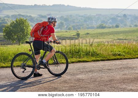 Ecotourism in Europe. Bicycle race in the country. Bicyclist traveler with backpack has adventure on european country road. Cyclist in white helmet and red t-shirt rides black mountain bike.