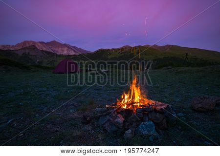 Tent And Burning Camp Fire At Dusk On The Mountains. Summer Adventures And Exploration In The Alps.