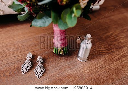 Elegant bridal earrings and perfume on the wooden floor next to the wedding bouquet. Close-up