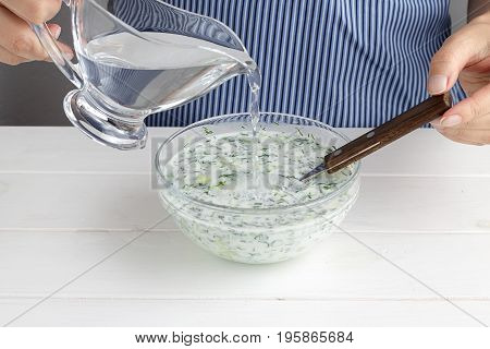 Step By Step Recipe For Tarator. Hands Pour Water Into A Bowl With Mixed Vegetables And Yogurt.