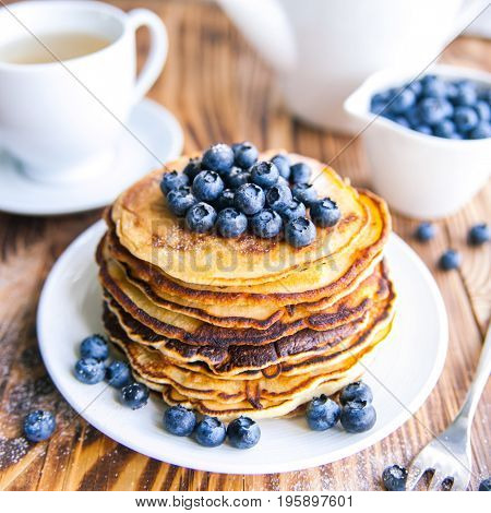 Pancakes healthy breakfast with blueberries, bog whortleberry, cup of green tea, cup of blueberries and teapot on brown wooden background