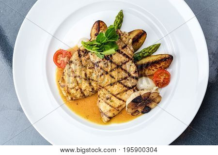Grilled chicken fillet served with french fries, asparagus and cherry tomatoes, decorated with grilled garlic and baby spinach
