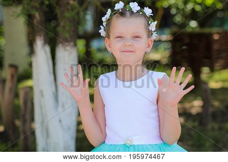 Cute little girl in a blue and white dress and wreath raises her hands she said - hello