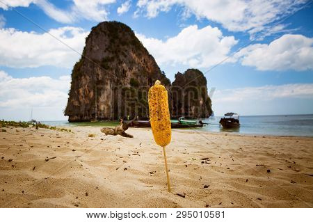 Ear Of Fried Corn On A Stick. Stuck In The Sand On A Tropical Beach. Close-up. Mountain, Sea And Blu