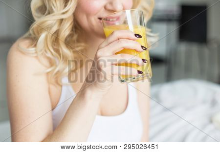 Attractive Blonde Woman Sits On The Edge Of The Bed At Home And Holds A Stocker With Orange Juice In