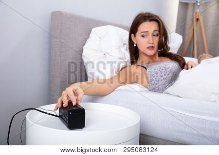 Young Woman In Bed Reaching For Snooze Button In Bedroom