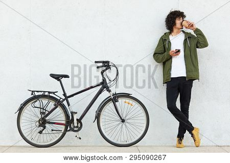 Studio Portrait Of Handsome Young Caucasian Male With Curly Hair And Little Beard, Drinking Coffee A