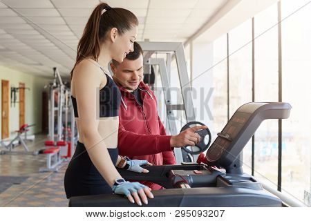 Cheerful Magnetic Young Lady Asking For Help Her Trainer, Trying To Use Running Machine, Listening T