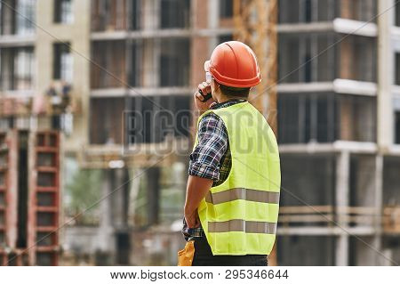 Construction Process. Back View Of Professional Young Builder In Working Uniform, Red Protective Hel