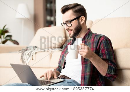 Young casual employee with mug of tea and laptop sitting by couch in living-room and working remotedly